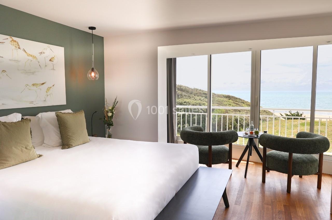 Chambre lumineuse avec lit double, fauteuils, table et vue sur la mer depuis une grande baie vitrée.