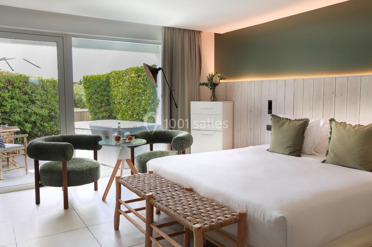 Chambre lumineuse avec lit double, fauteuils verts, table basse, et accès à une terrasse avec jacuzzi et végétation.