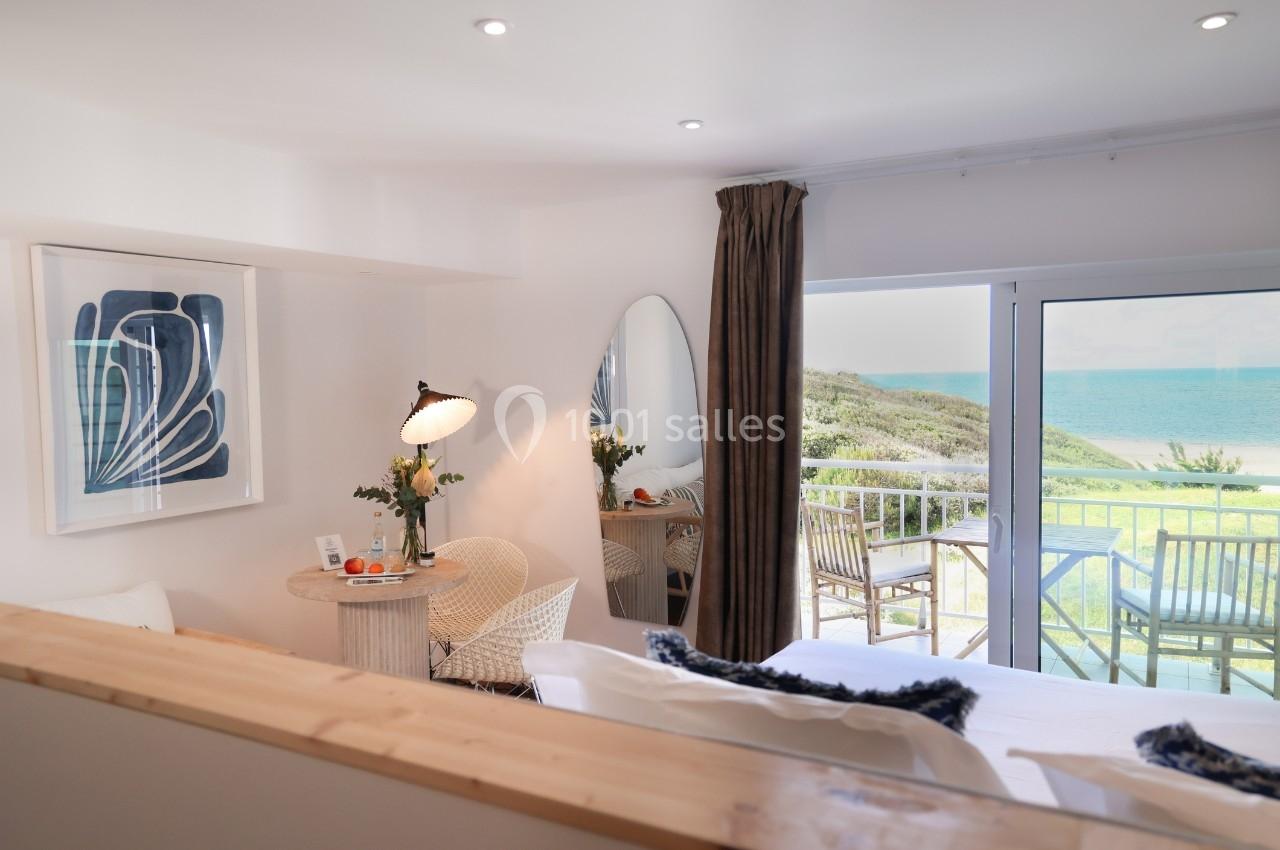 Chambre lumineuse avec lit, table, fauteuil, balcon donnant sur la mer et décoration moderne.