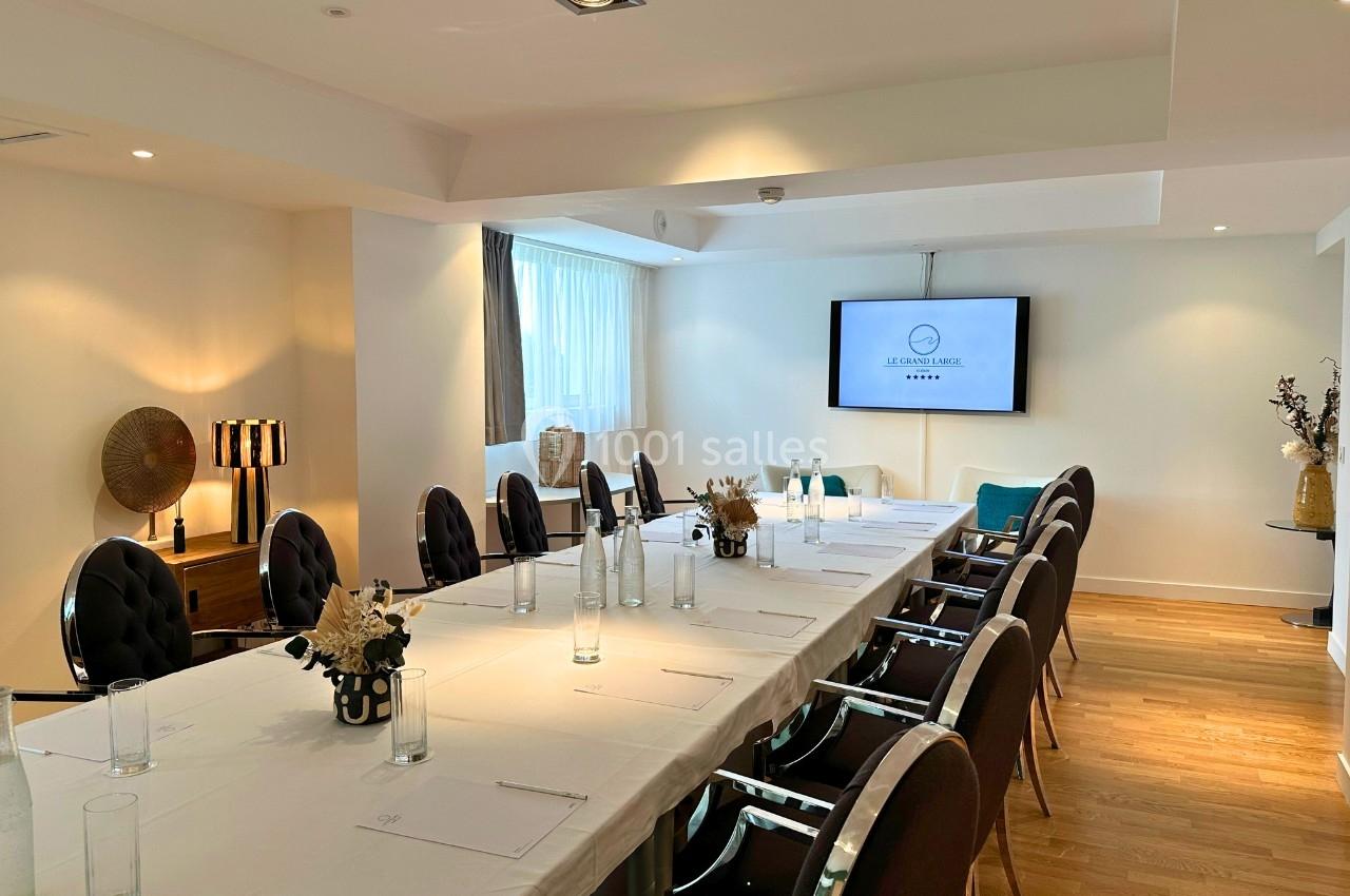 Salle de réunion lumineuse avec une grande table rectangulaire, chaises noires, écran mural et décor sobre.