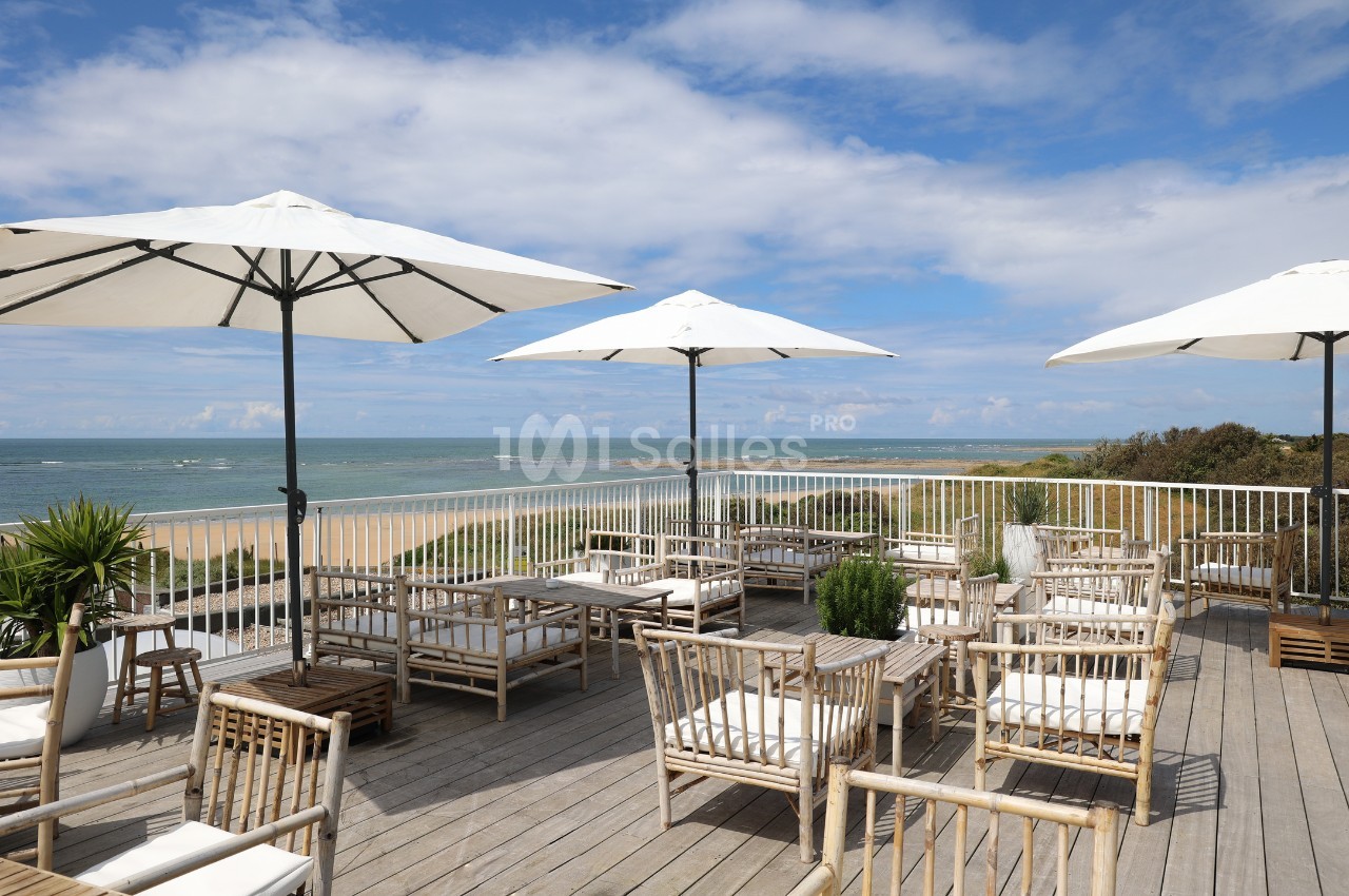 Terrasse en bois avec mobilier en bambou et parasols, offrant une vue sur la plage et la mer sous un ciel partiellement…