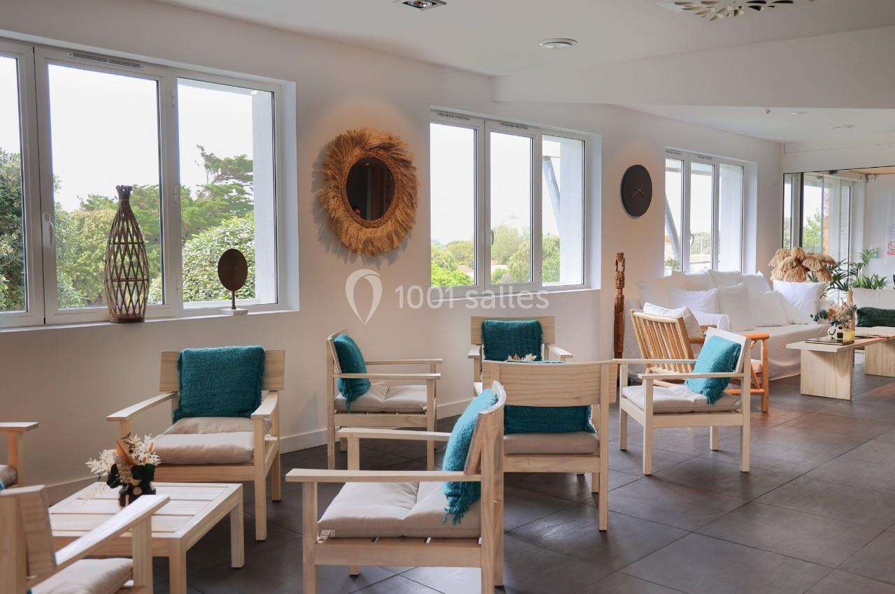 Espace lumineux avec fauteuils en bois, coussins bleus, grandes fenêtres et décoration naturelle.