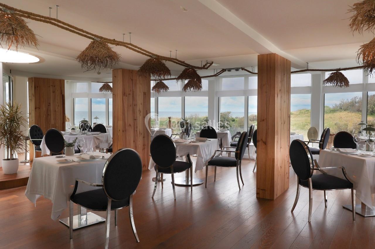 Salle de restaurant lumineuse avec tables dressées, chaises noires et vue sur un paysage naturel à travers de grandes baies…