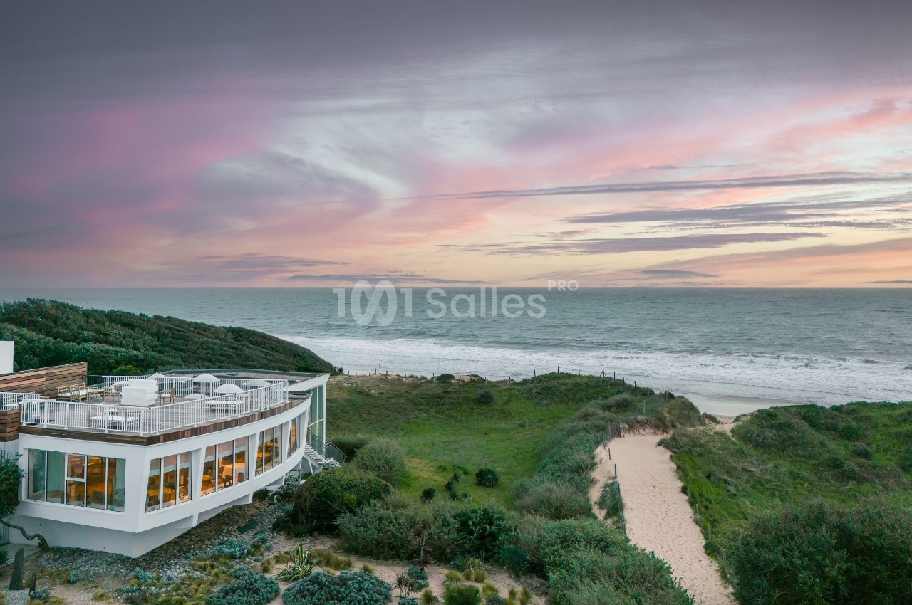 Vue d'une maison moderne avec terrasse, située près d'un sentier menant à une plage, sous un ciel coloré au coucher du…