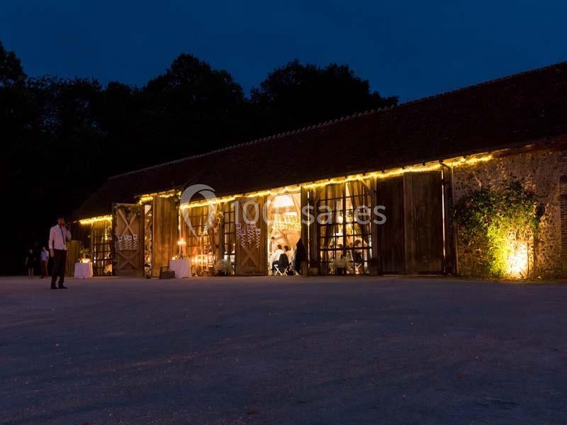 Grange en soirée éclairée par des guirlandes lumineuses, avec des portes ouvertes laissant entrevoir un événement à l…