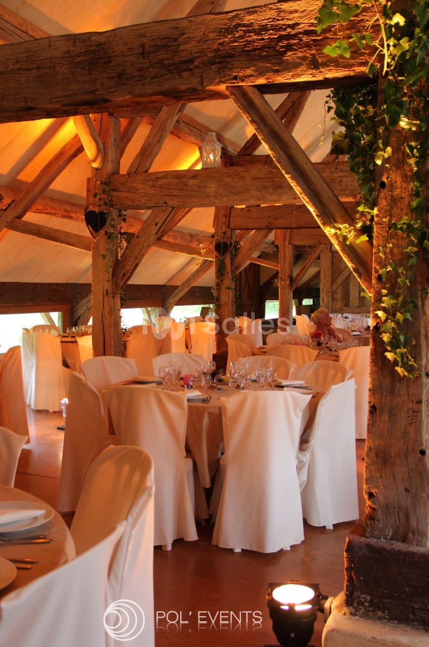 Salle de réception rustique avec poutres apparentes, tables rondes dressées et chaises couvertes de housses blanches.