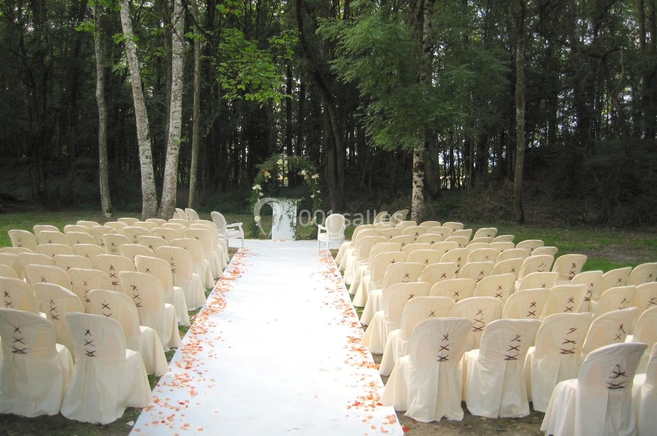 Allée centrale bordée de chaises blanches décorées, menant à un autel floral en plein air dans une forêt.