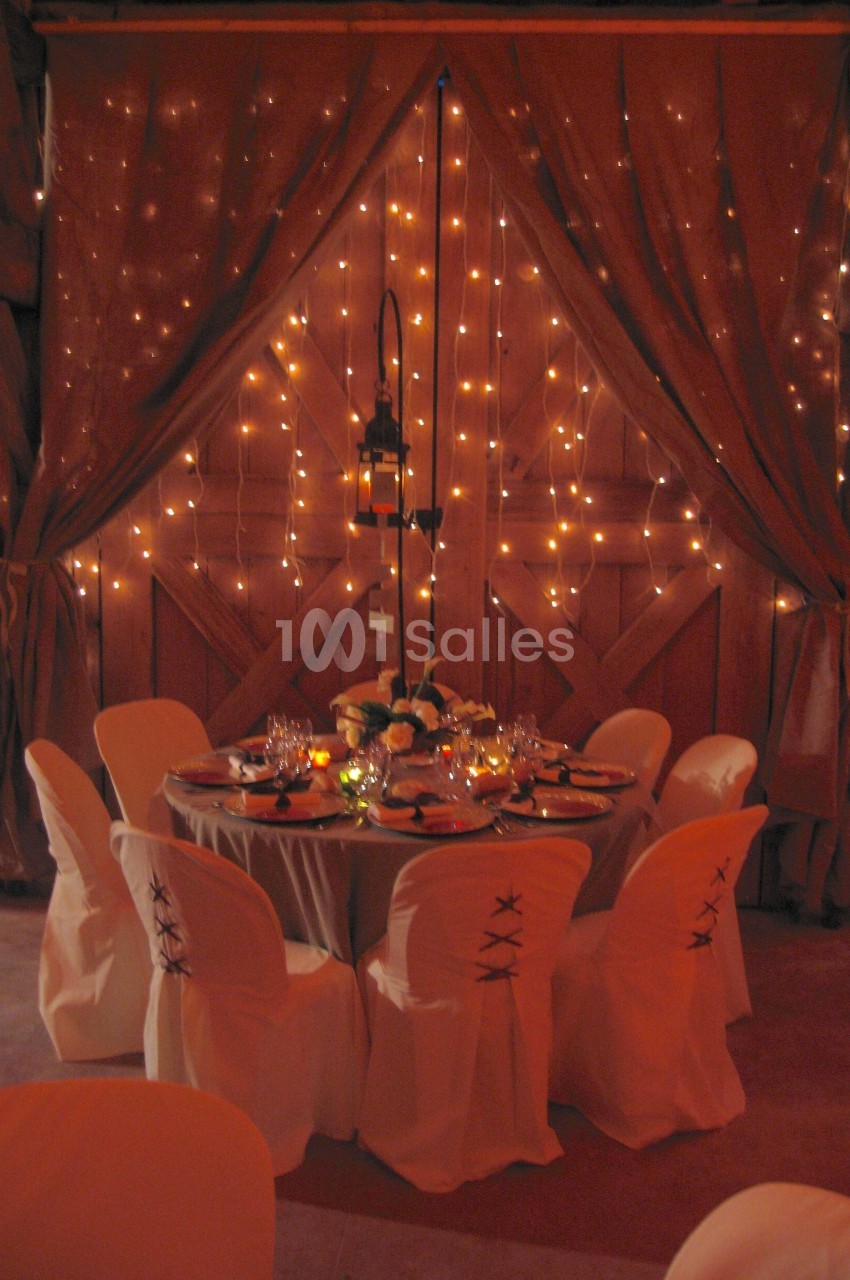 Table ronde décorée pour un dîner, entourée de chaises blanches, devant un rideau illuminé de guirlandes lumineuses.