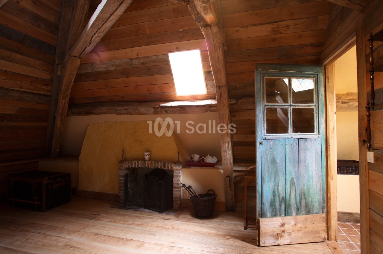 Grenier aménagé avec poutres en bois apparentes, cheminée, coffre en bois, et porte ancienne entrouverte.