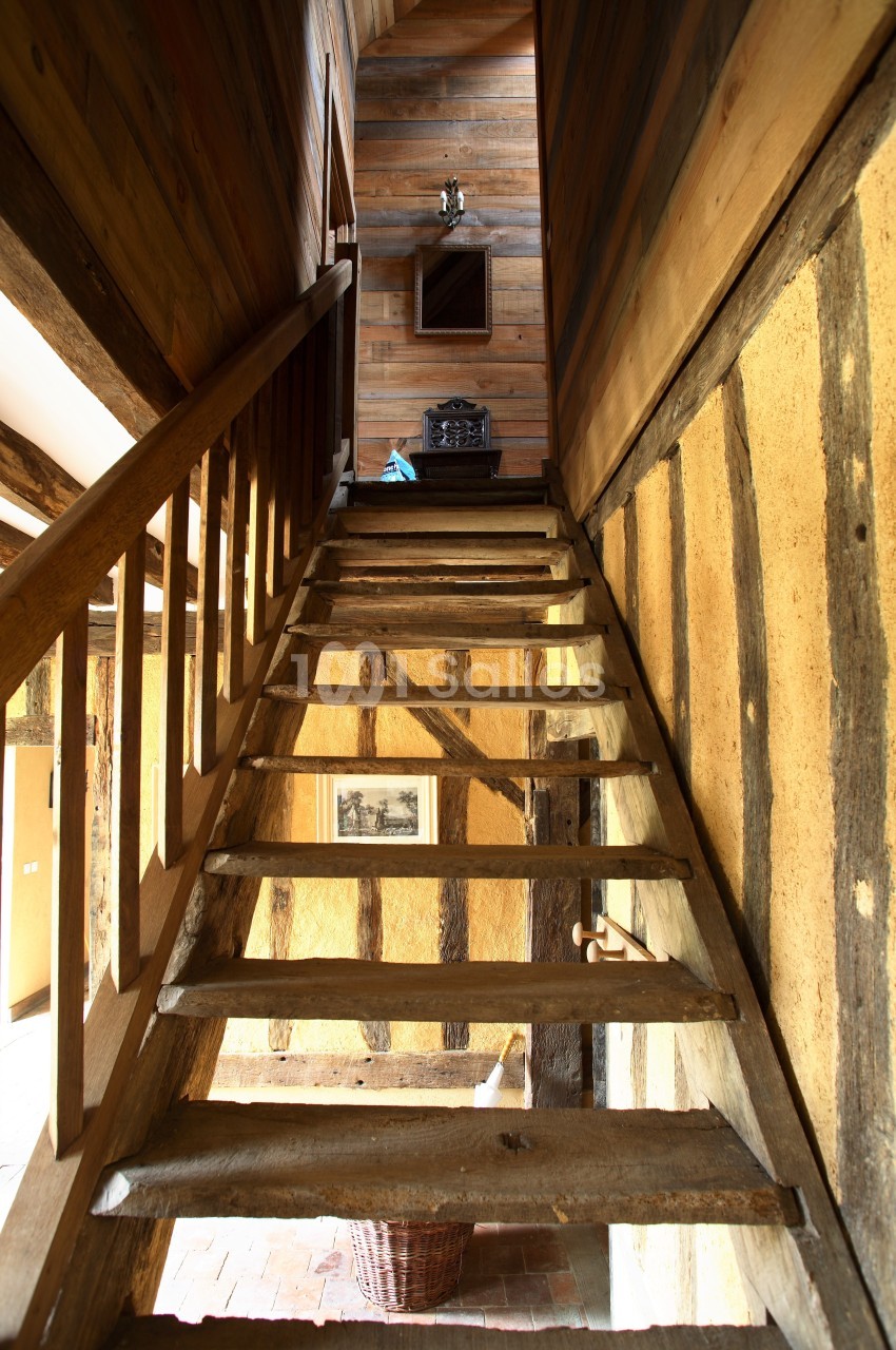 Escalier en bois rustique montant vers un étage, entouré de murs en bois et torchis dans une maison ancienne.