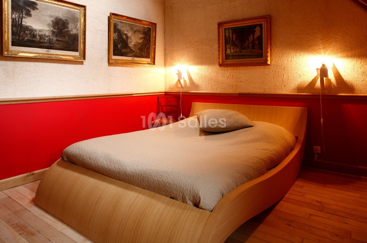 Lit double avec couverture beige dans une chambre aux murs rouges et blancs, éclairée par deux appliques murales.