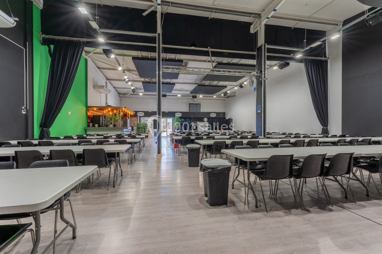 Salle spacieuse avec des tables et chaises alignées, éclairage artificiel et rideaux noirs encadrant la scène.