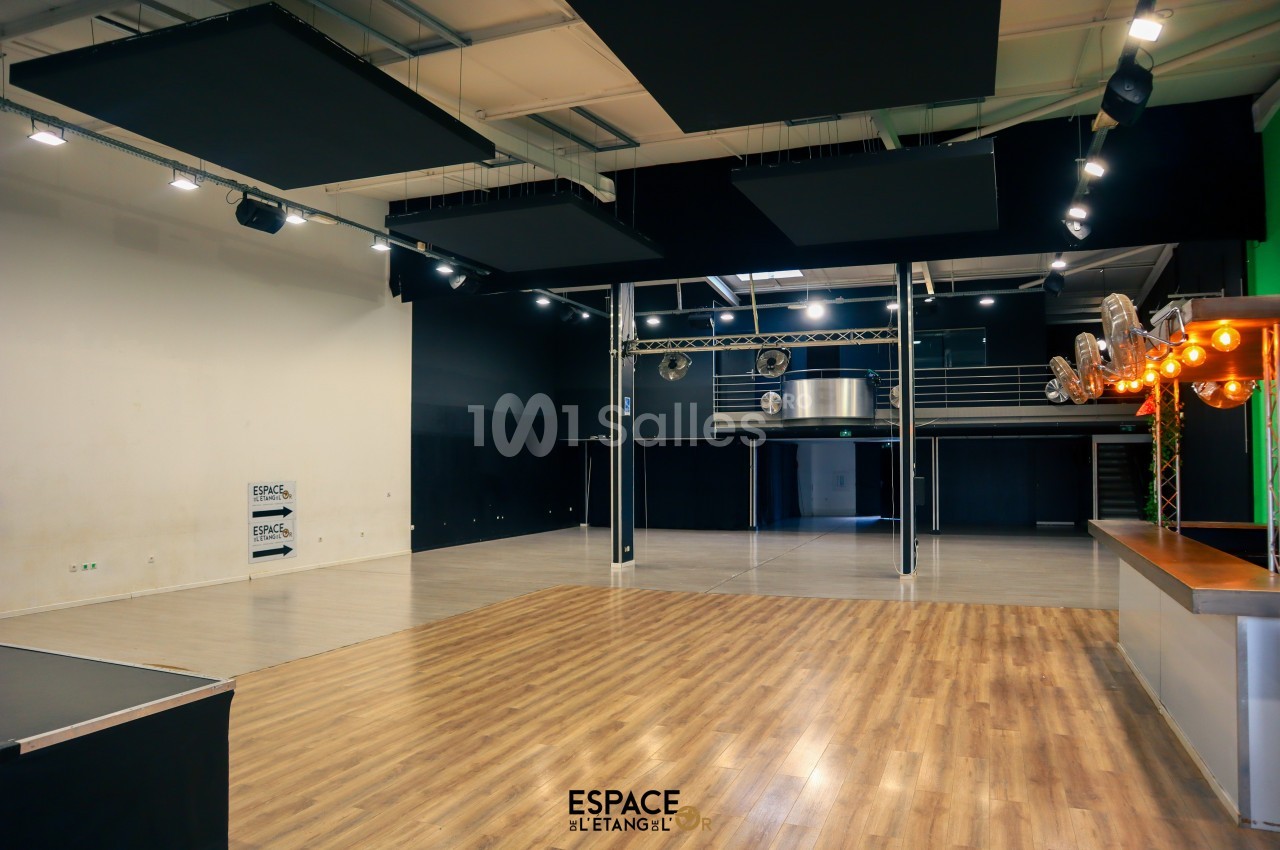Salle polyvalente avec parquet, espace scénique, mezzanine métallique et éclairage suspendu.