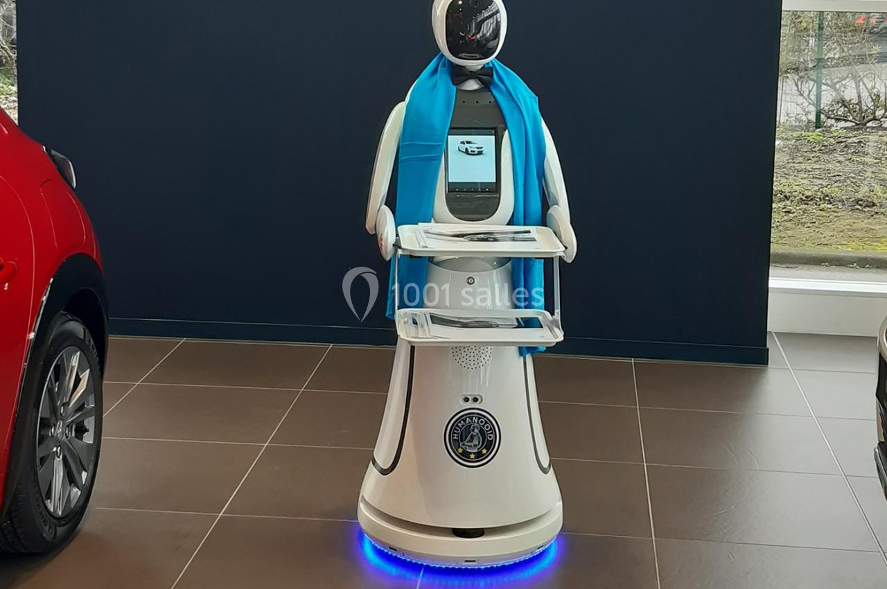 Robot blanc avec écran intégré, écharpe bleue et plateaux, exposé dans un espace intérieur avec des voitures en arrière-plan.