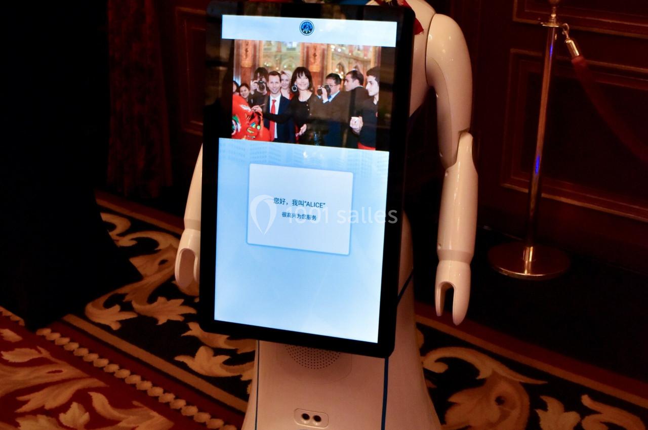 Robot humanoïde blanc avec écran affichant un visage animé, exposé dans une pièce ornée de rideaux rouges et tapis.