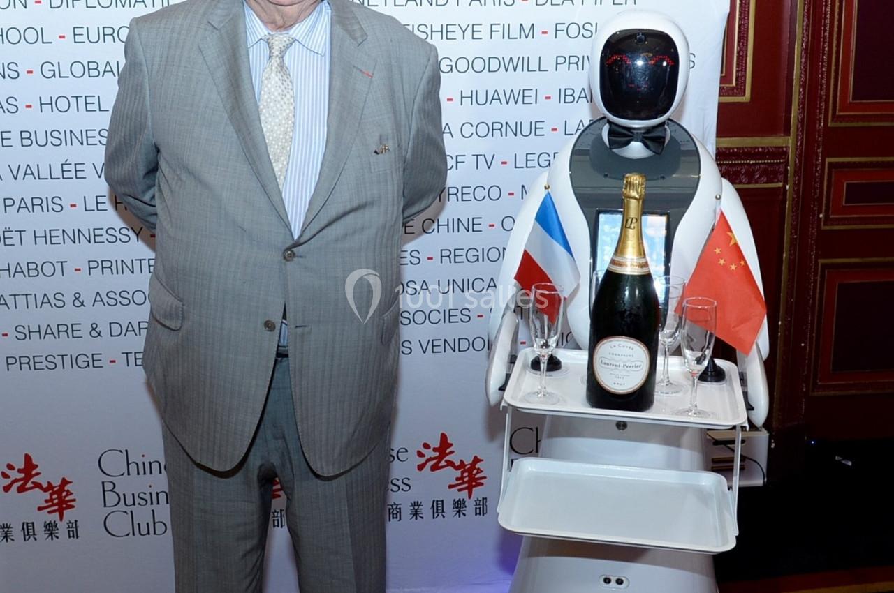 Un homme en costume pose à côté d'un robot portant une bouteille de champagne et des drapeaux français et chinois.
