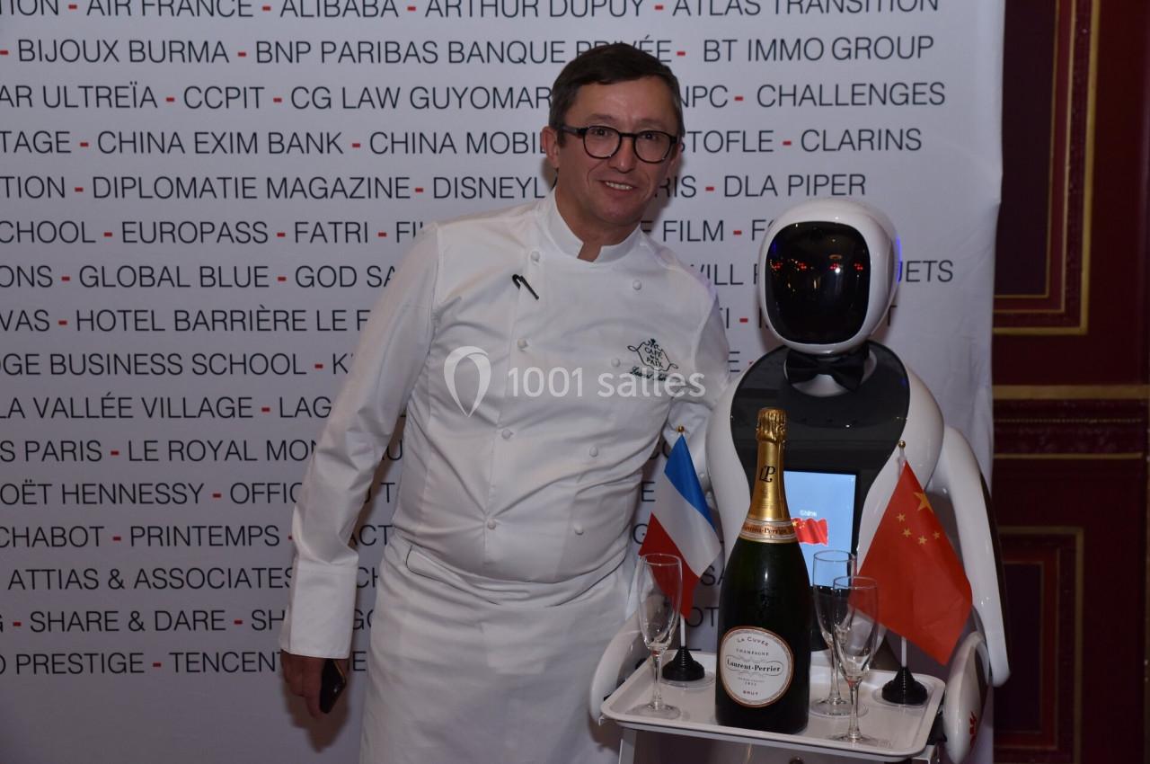 Un chef en tenue blanche pose avec un robot portant des drapeaux, une bouteille de champagne et des verres.