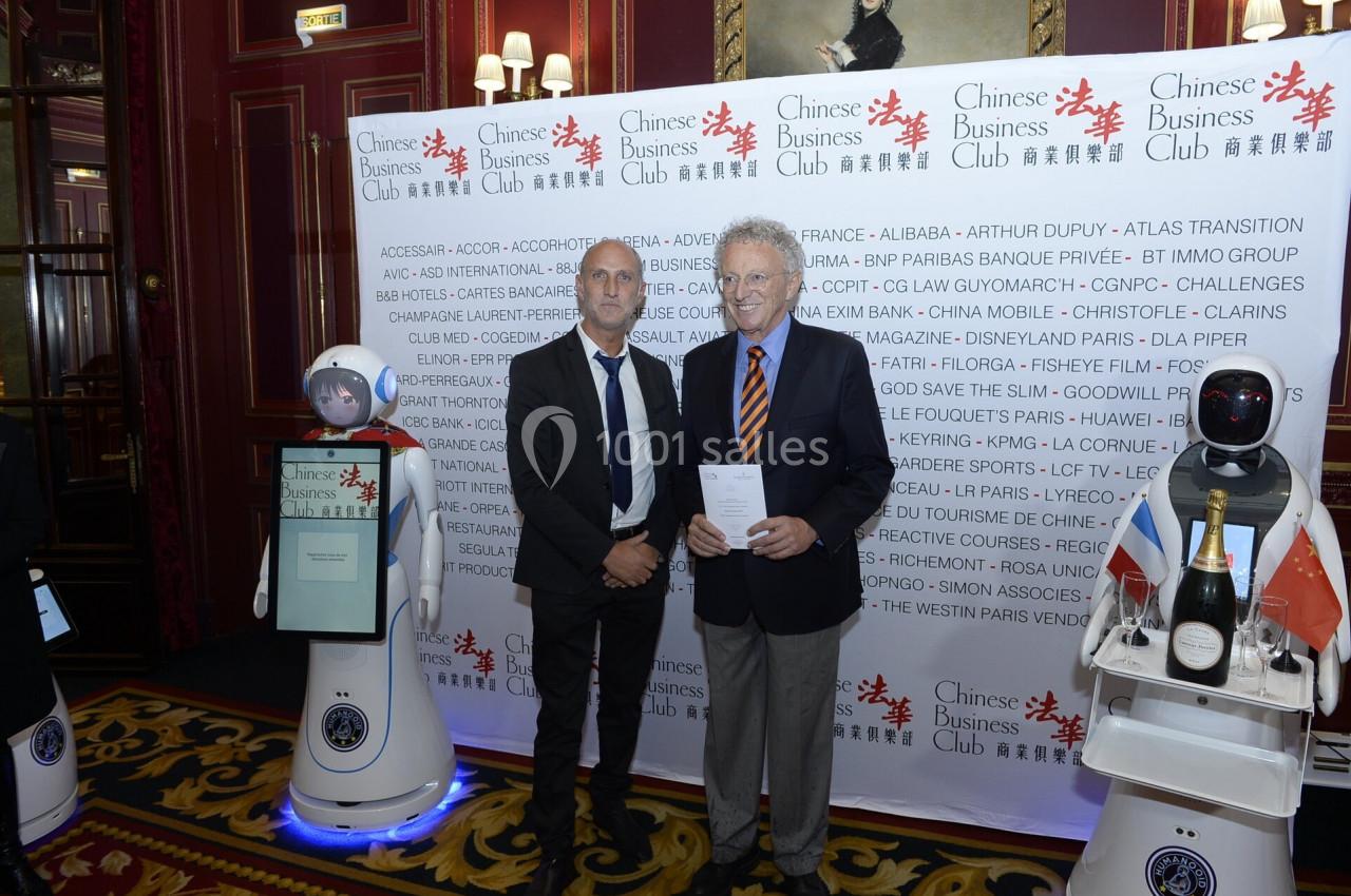 Deux hommes posent devant un panneau promotionnel dans un cadre élégant, entourés de robots et de décorations.