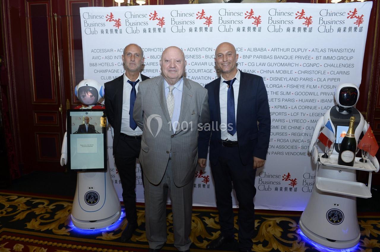 Trois hommes posent devant un mur avec des logos, entourés de deux robots interactifs sur un tapis rouge.