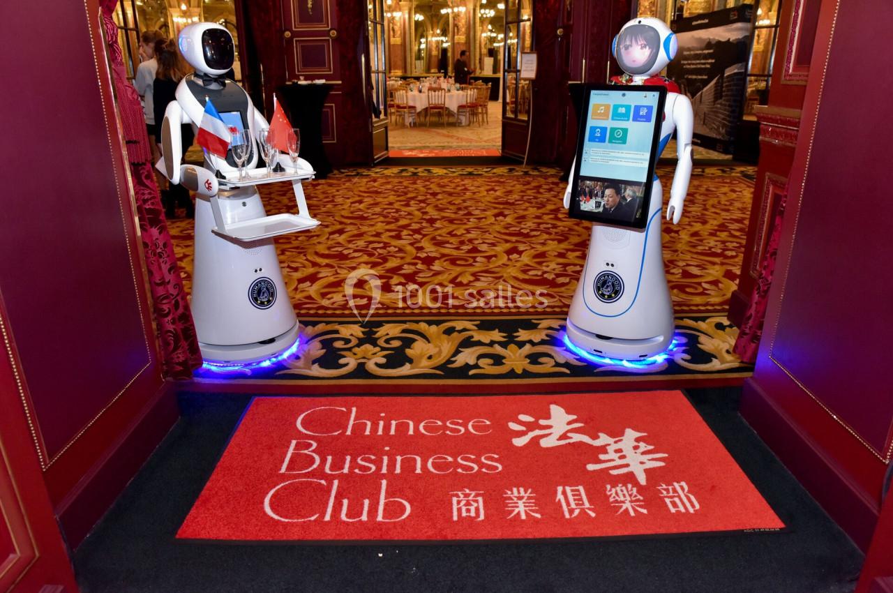 Deux robots humanoïdes accueillent les visiteurs à l'entrée d'un événement, sur un tapis rouge avec des inscriptions en…