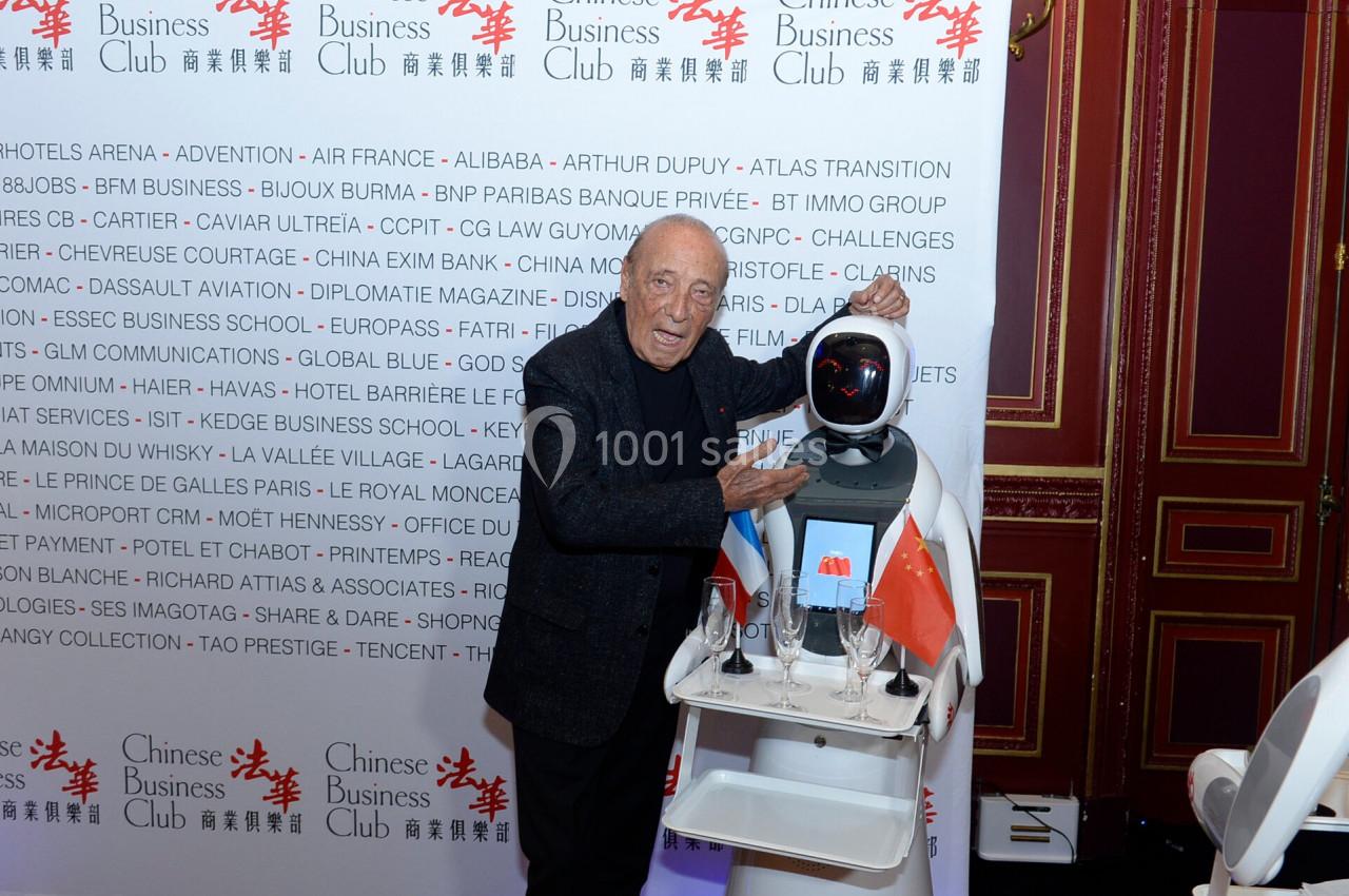 Un homme pose avec un robot humanoïde équipé d'un écran, devant un mur avec des logos d'entreprises.