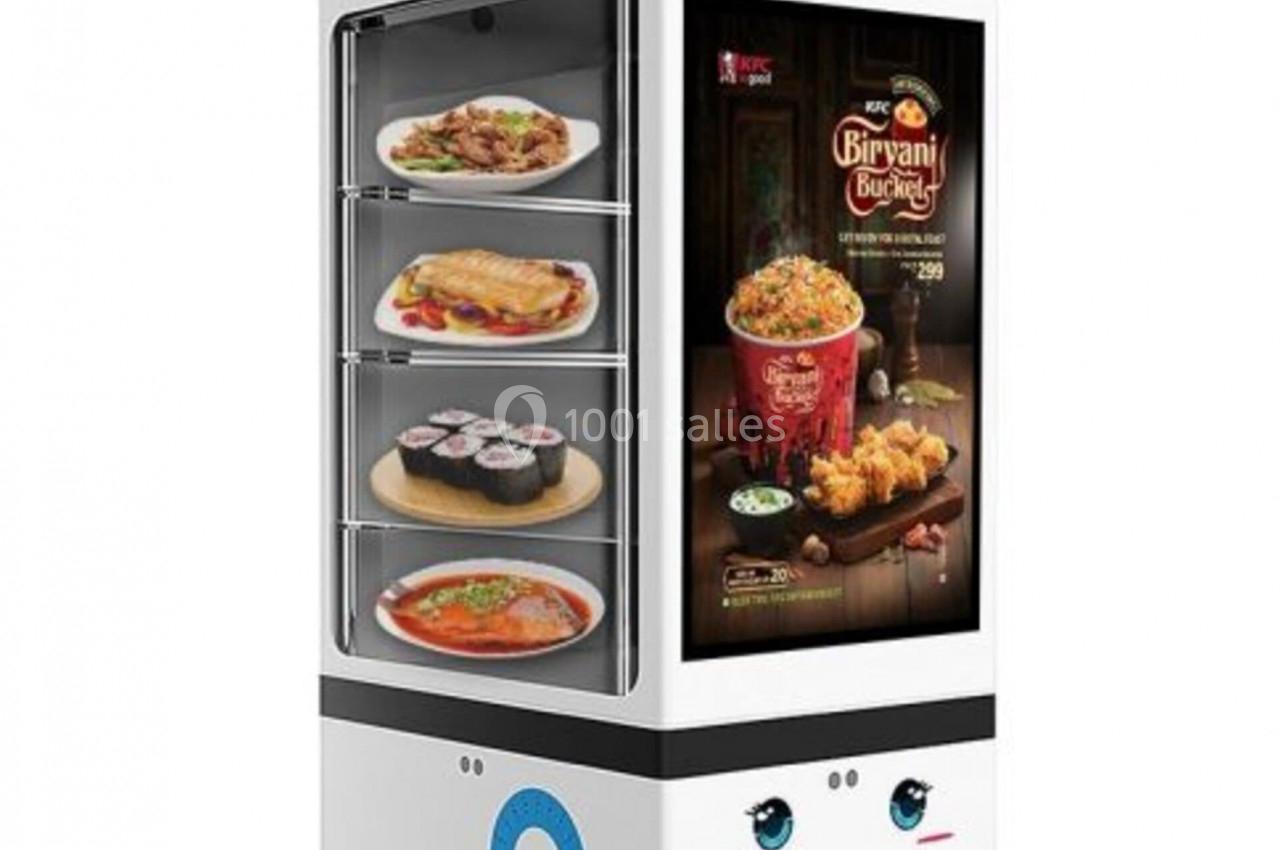 Un robot distributeur affichant des plats variés dans des compartiments vitrés et un écran publicitaire.