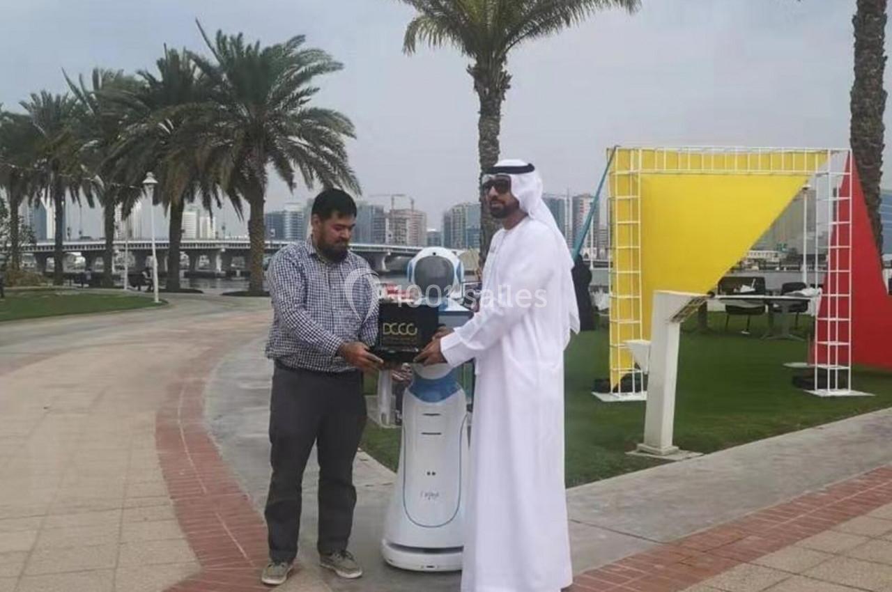 Deux hommes interagissent avec un robot dans un parc bordé de palmiers, sous un ciel nuageux.