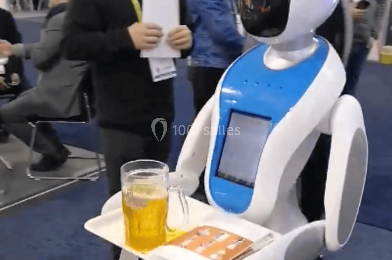 Un robot serveur blanc et bleu portant une chope de bière et des plateaux, entouré de personnes dans un salon professionnel.