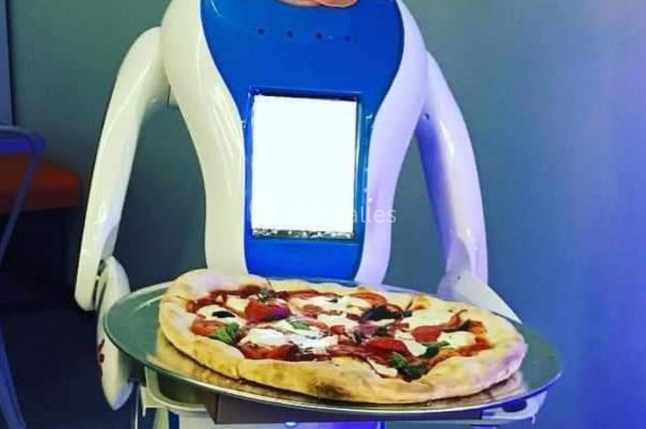 Robot humanoïde bleu et blanc servant une pizza sur un plateau, avec des assiettes et des couverts en dessous.