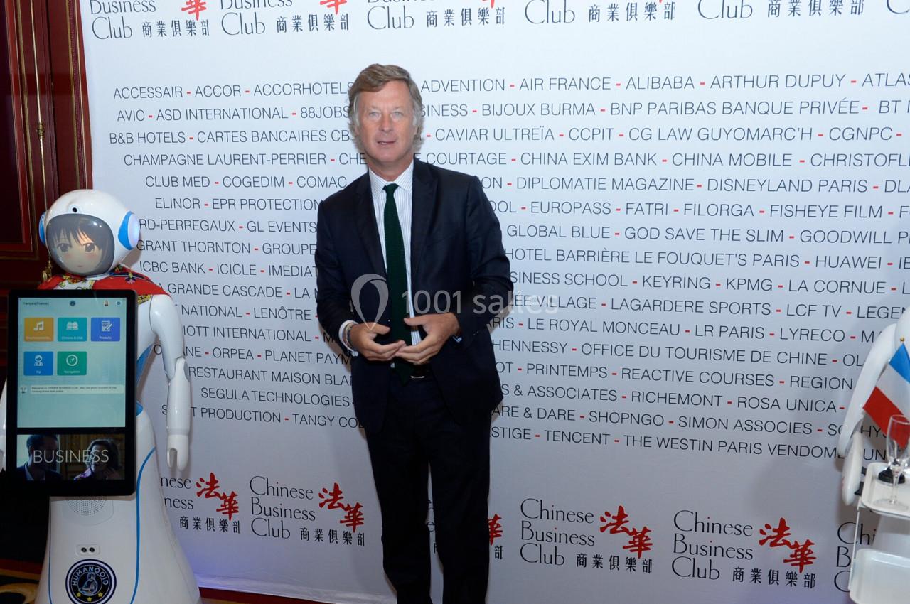 Un homme en costume pose debout devant un mur couvert de logos et de textes, avec un robot humanoïde à ses côtés.