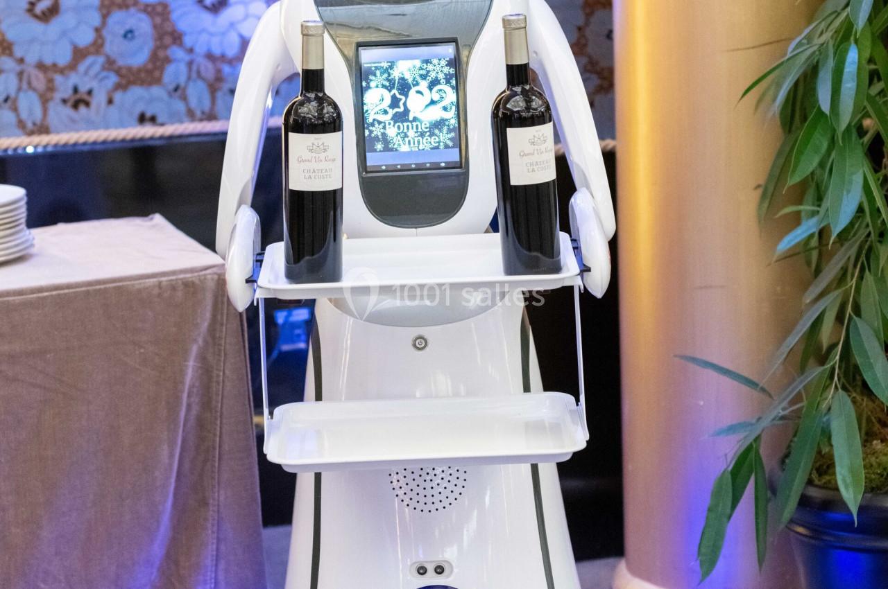 Robot blanc équipé d'un écran et portant deux bouteilles de vin sur un plateau, dans un environnement intérieur élégant.