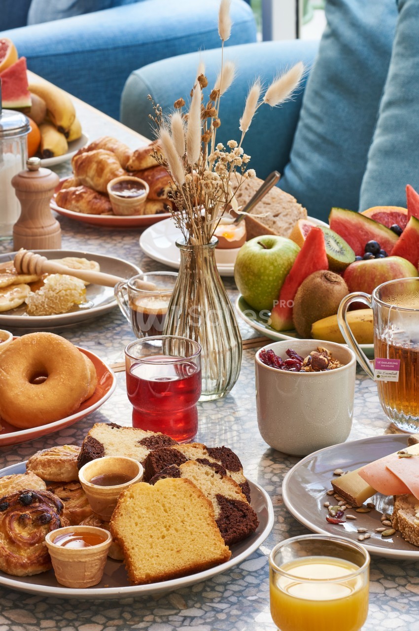 Petit-déjeuner varié avec viennoiseries, fruits frais, boissons et pain sur une table lumineuse et conviviale.