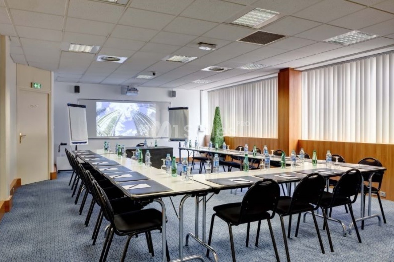 Salle de réunion lumineuse avec tables disposées en U, chaises noires, écran de projection et fournitures de bureau.