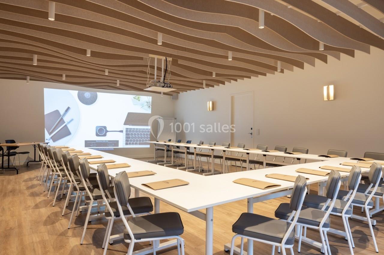 Salle de réunion moderne avec tables disposées en U, chaises alignées et projection d'une présentation sur un écran.
