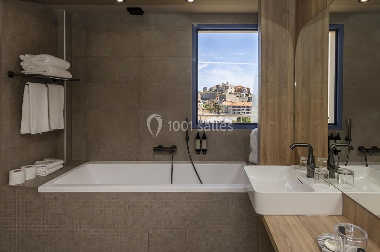 Salle de bain moderne avec baignoire, lavabo, serviettes pliées et fenêtre offrant une vue sur des bâtiments extérieurs.