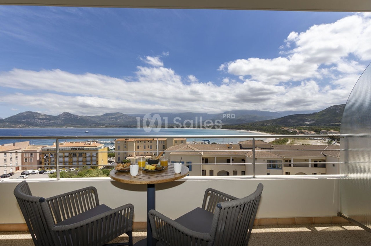 Vue depuis un balcon avec table et chaises, donnant sur une baie, des bâtiments et des montagnes sous un ciel partiellement…