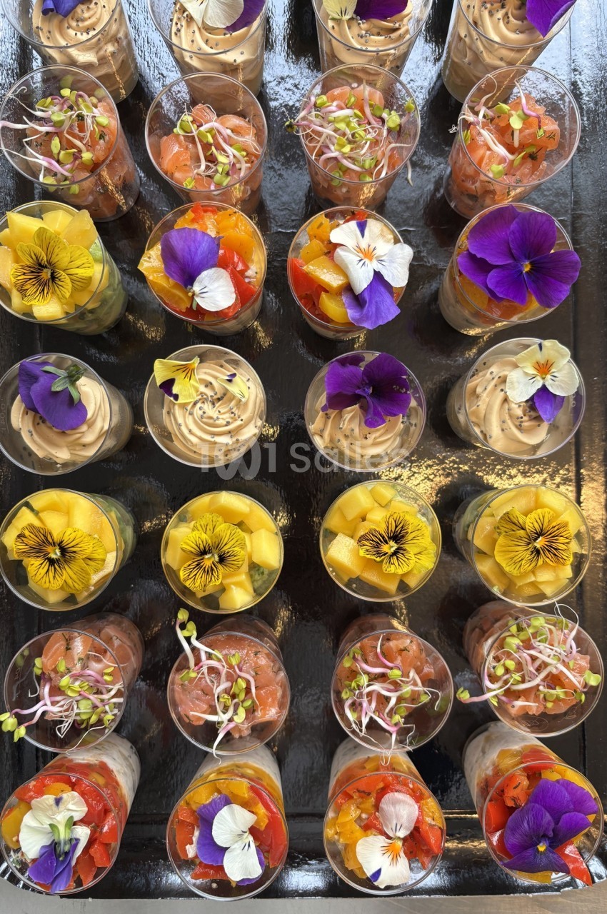 Plateau de verrines colorées garnies de fleurs comestibles, mousses et légumes, disposées en rangées.