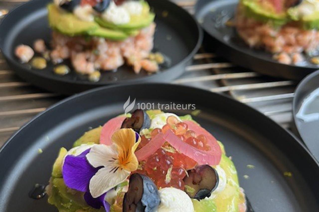 Tartare de saumon surmonté d'avocat, fromage frais, fleurs comestibles et fruits, présenté sur une assiette noire.