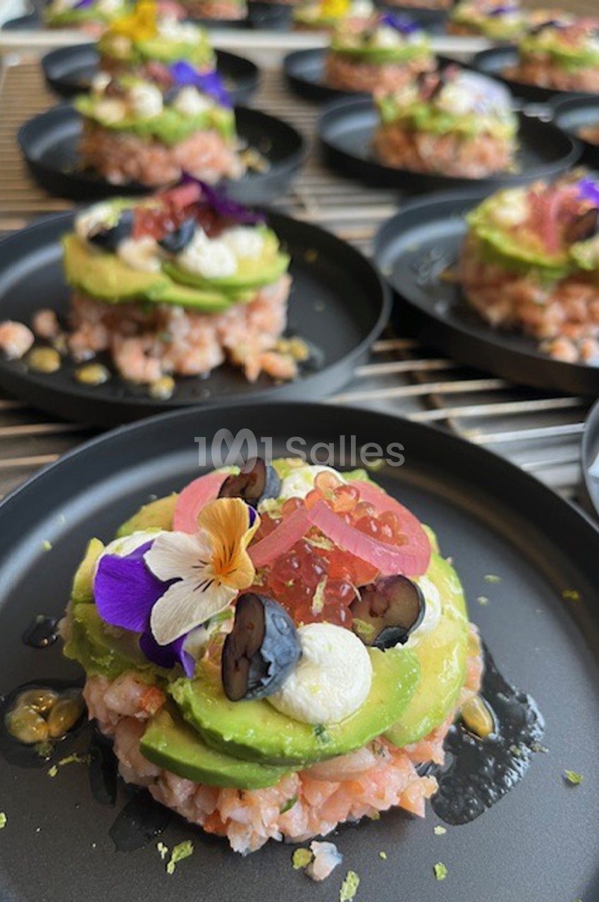 Tartare de saumon surmonté d'avocat, fromage frais, fleurs comestibles et fruits, présenté sur une assiette noire.