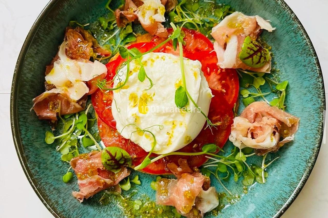 Assiette garnie de burrata, tranches de tomate, jambon cru, herbes fraîches et filet d'huile sur un fond clair.