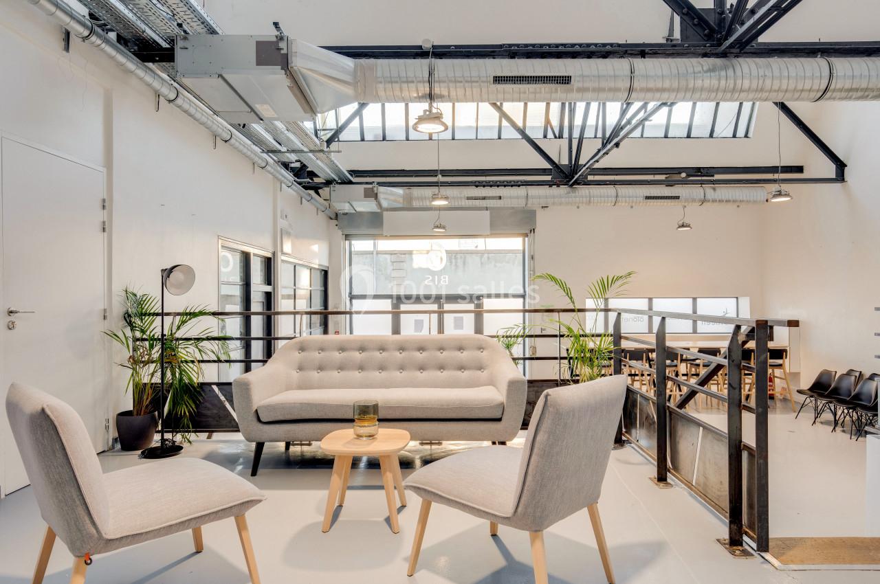 Espace lumineux avec canapé gris, fauteuils assortis, plantes et grande verrière dans un intérieur industriel.