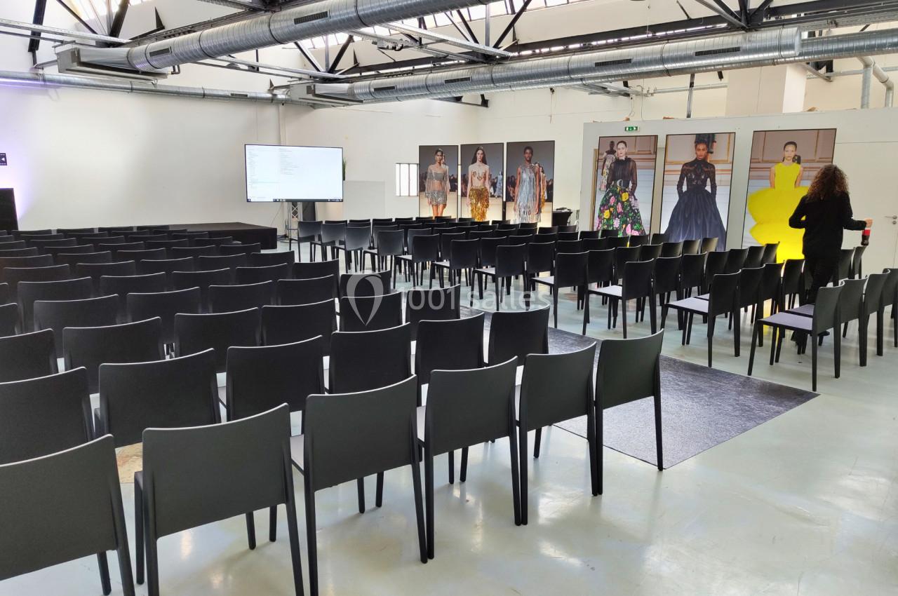 Salle de conférence vide avec des rangées de chaises noires, un écran de projection et des affiches de mode en arrière-plan.