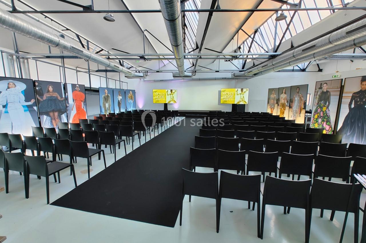 Salle d'événement avec rangées de chaises noires, podium central et écrans affichant des visuels de mode.