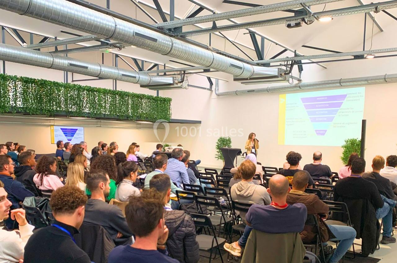 Conférencière présentant un diaporama devant un public assis dans une salle lumineuse avec des plantes décoratives.
