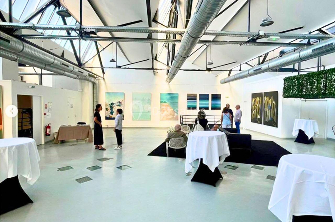 Salle d'exposition lumineuse avec des œuvres d'art accrochées aux murs, quelques personnes discutent debout.