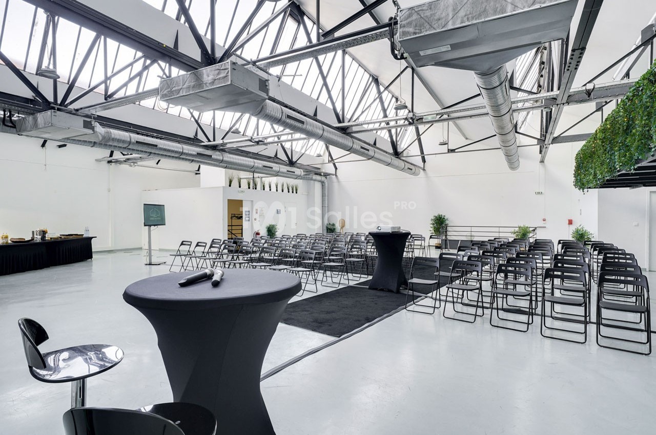Salle lumineuse avec plafond industriel, rangées de chaises, tables hautes et buffet en arrière-plan.