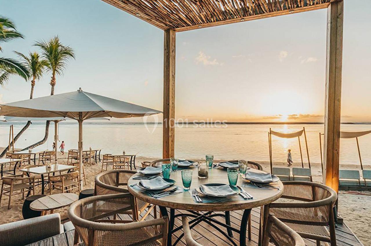 Terrasse en bord de plage avec table dressée, chaises en bois, parasol et vue sur un coucher de soleil au-dessus de l'océan.