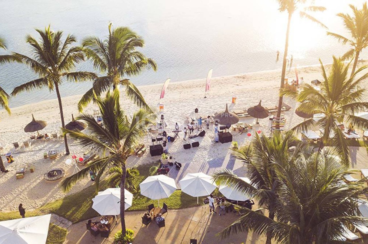 Vue aérienne d'une plage tropicale avec palmiers, parasols blancs, cabanes en chaume et activités en bord de mer.