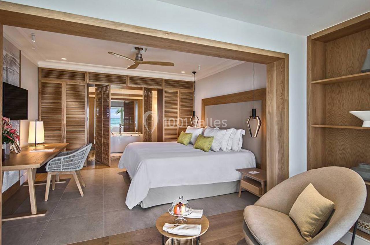 Chambre lumineuse avec lit double, bureau, fauteuil, étagères en bois et vue sur une salle de bain ouverte.