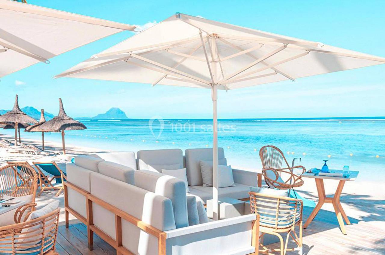 Terrasse en bord de mer avec parasols, mobilier en bois et vue sur une plage de sable et une eau turquoise.